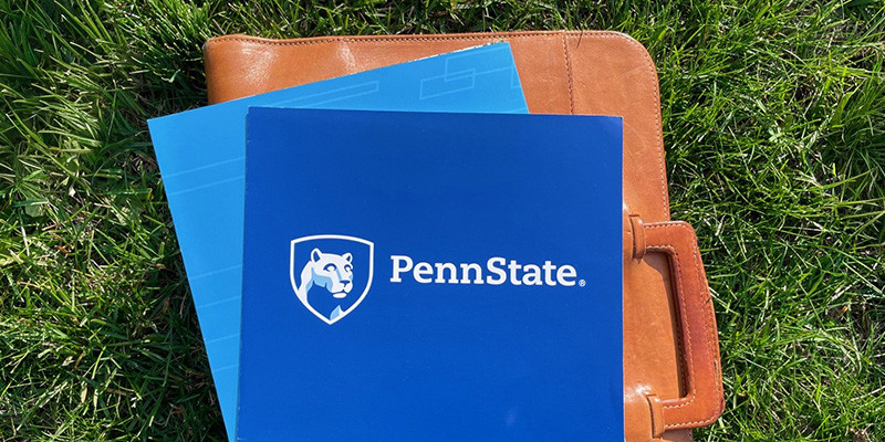 Penn State mark printed on a folder stacked with a briefcase in the grass, viewed from above
